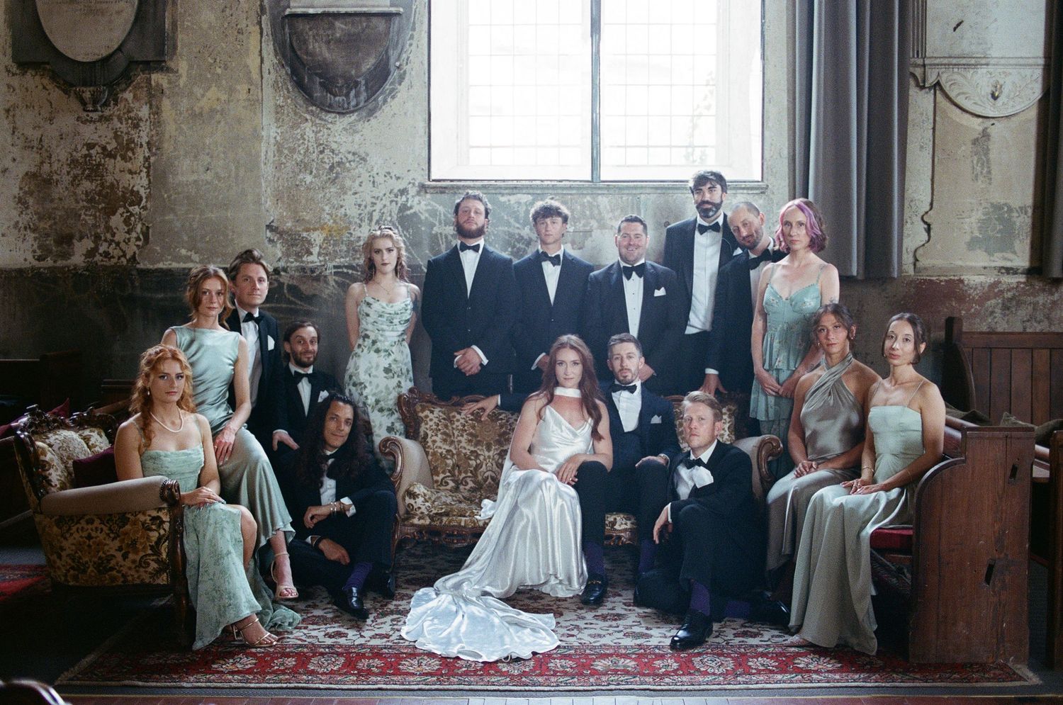 Large wedding party poses formally with bride and groom in a rustic historic venue interior.