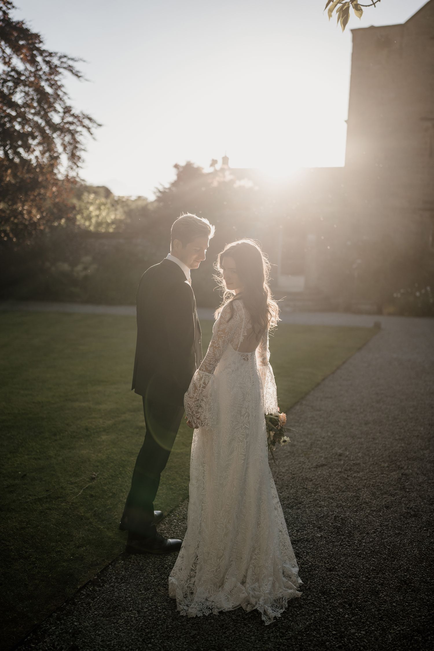 Bride and groom share intimate moment backlit by golden sunset outside historic stone wedding venue.