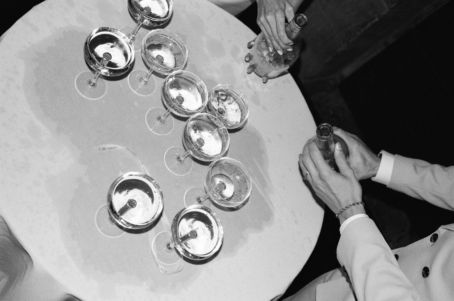 Black and white overhead view of wine glasses arranged on a round table with hands reaching for drinks.