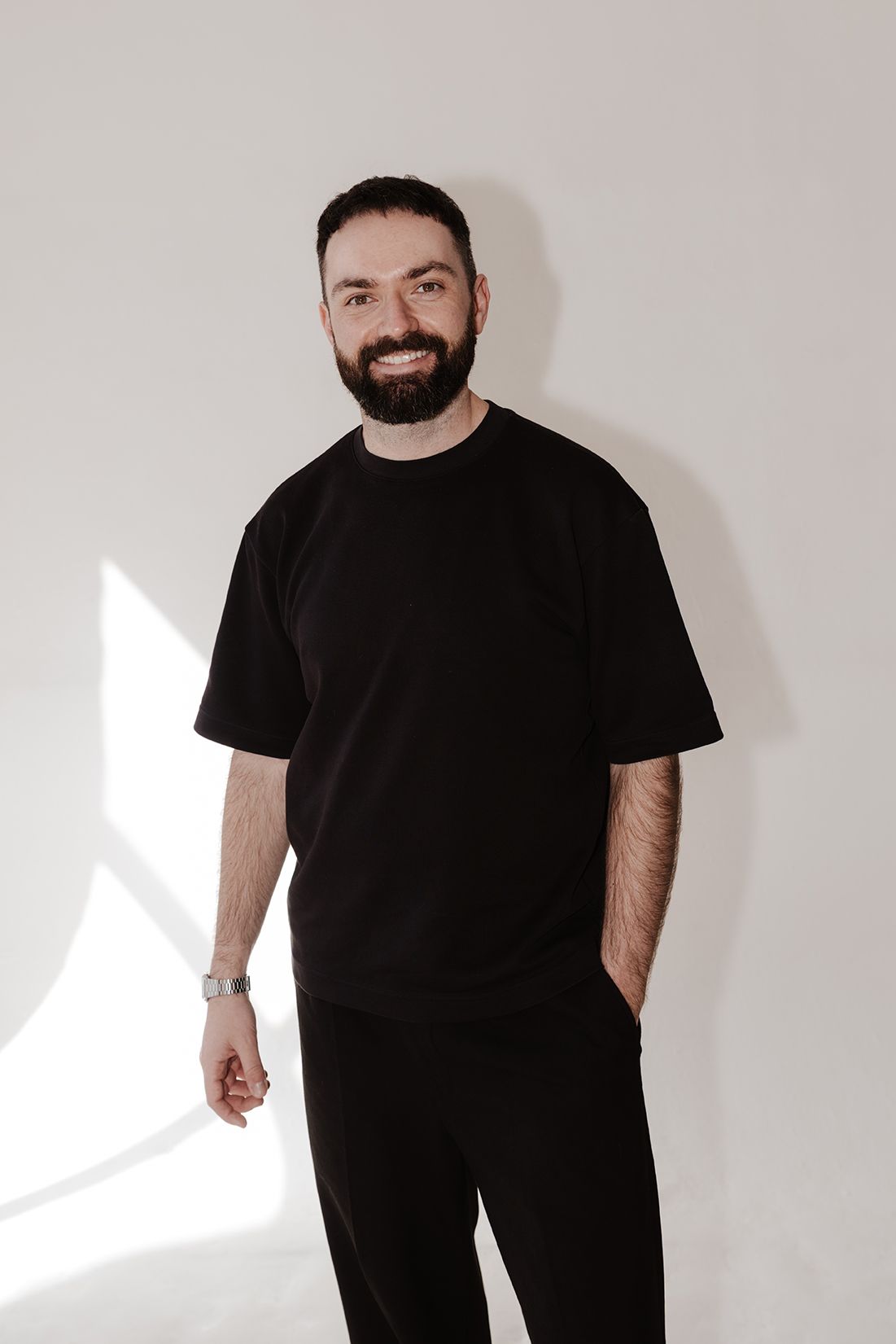 Smiling bearded man in black oversized t-shirt posing against white wall.