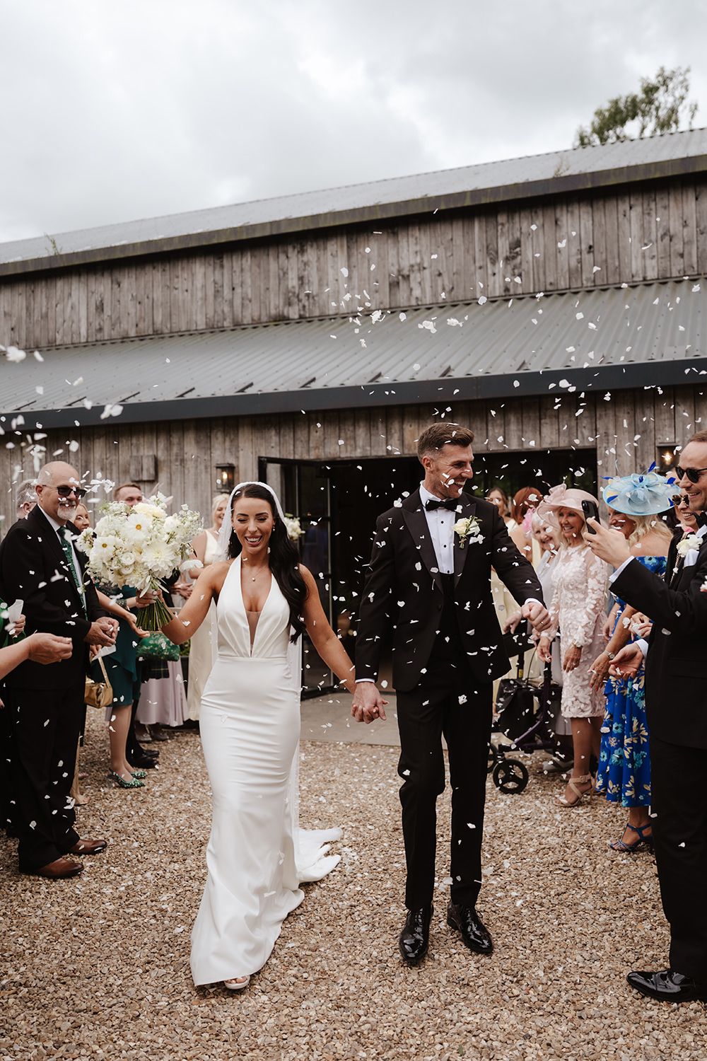 Wedding guests celebrate by throwing confetti at a rustic outdoor venue with wooden fence backdrop.