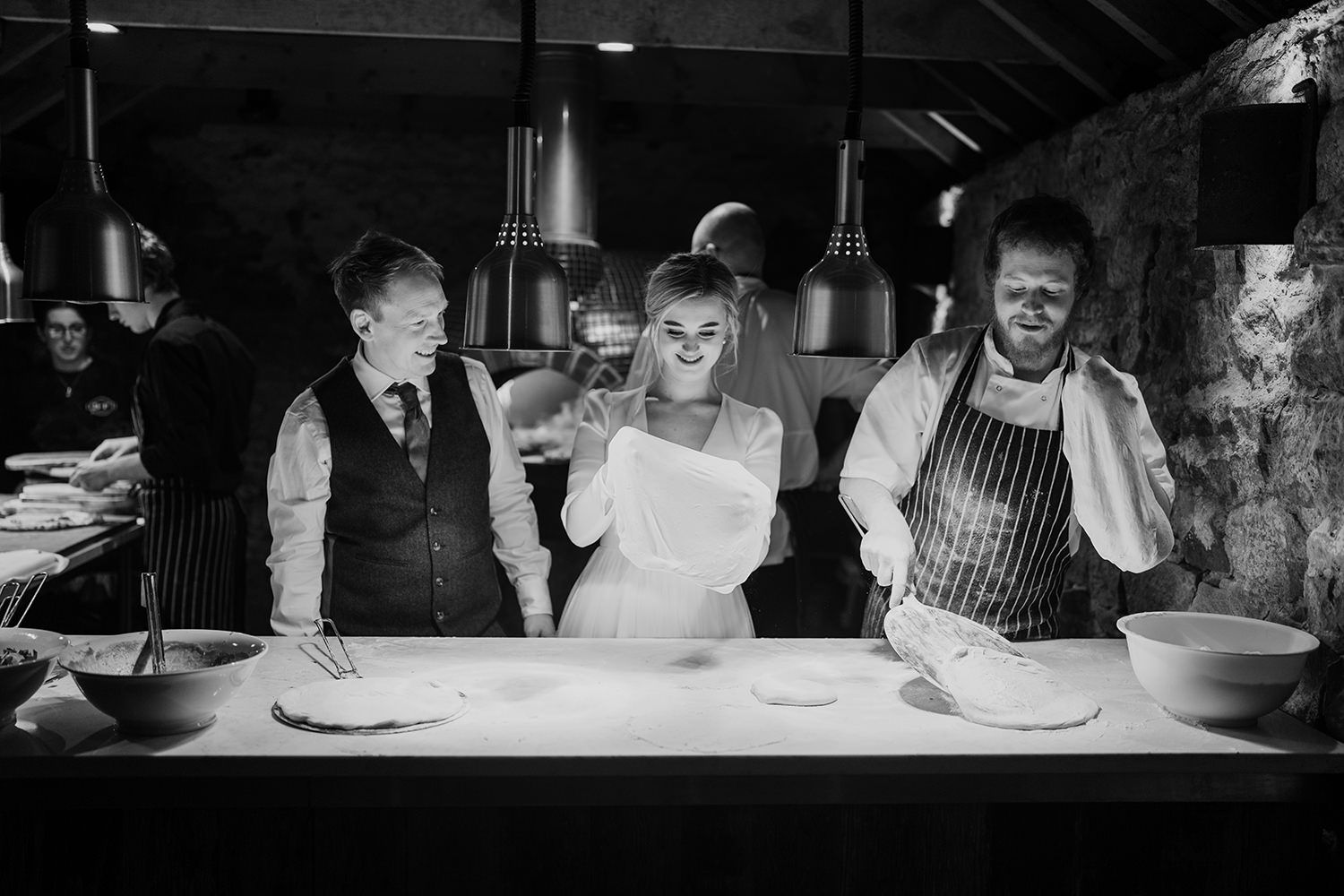 Bride and groom stood being shown how to make pizza on a white table.