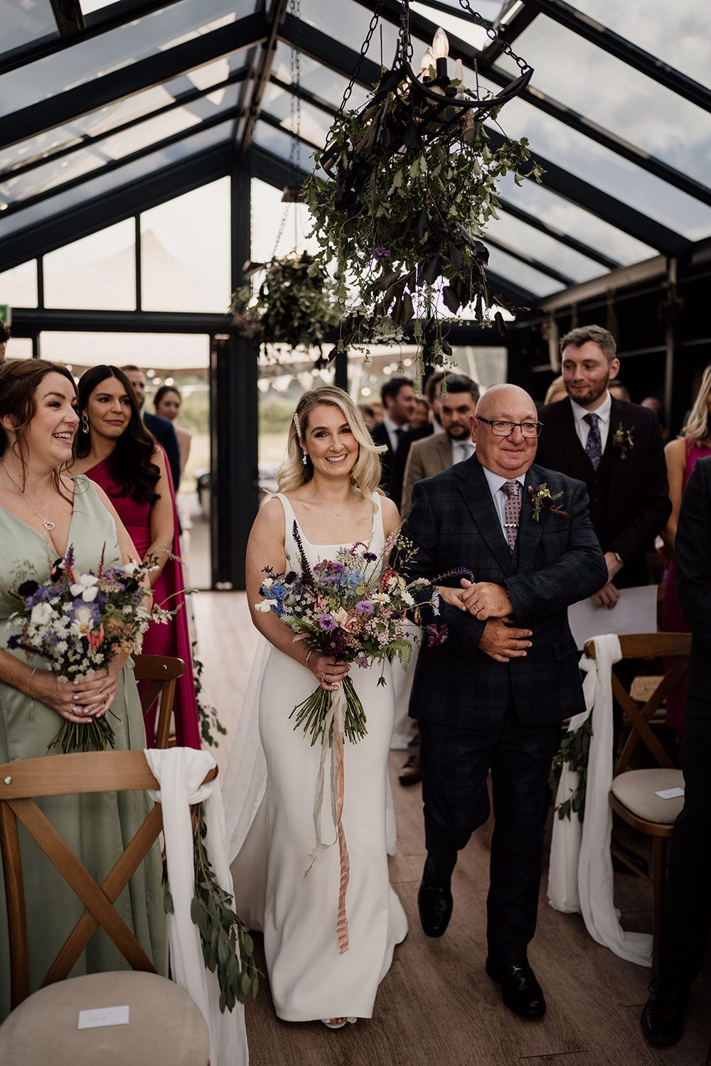 Bride and her dad walking down the wedding aisle, smiling, holding hands surrounded by wedding guests.