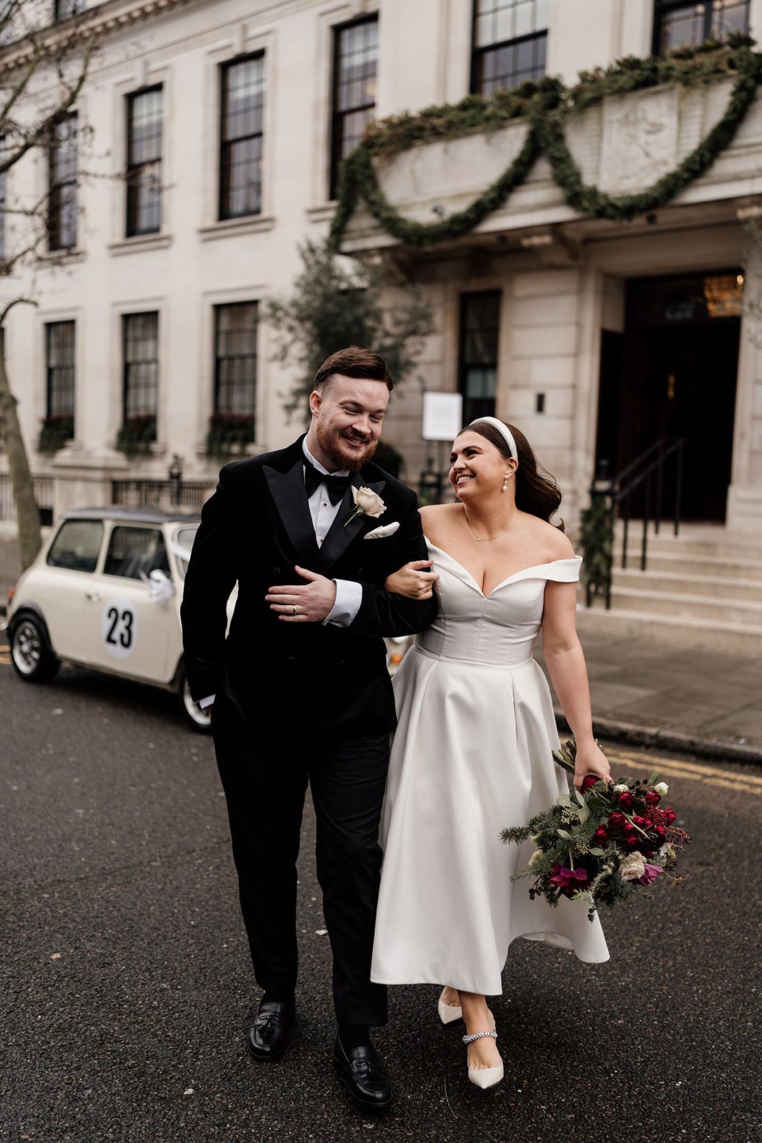 A couple walks arm in arm after their wedding ceremony, with a classic white dress and black tuxedo against a vintage building.