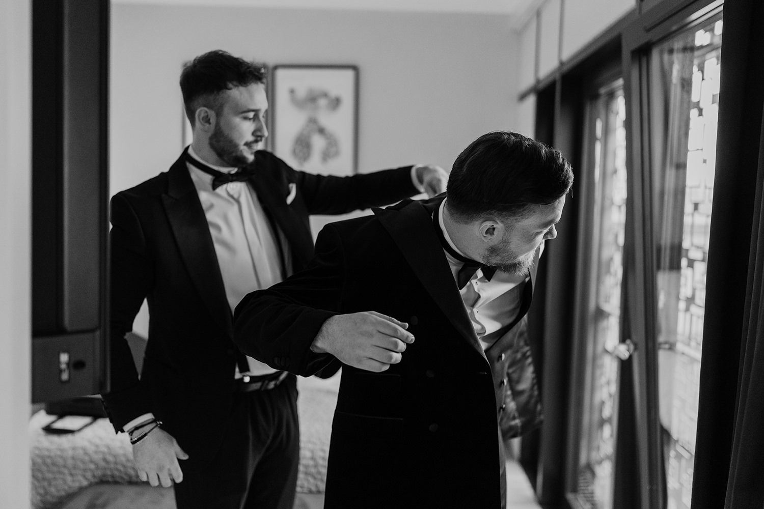A groomsman helps adjust the suit jacket of another in a candid black and white getting ready moment.