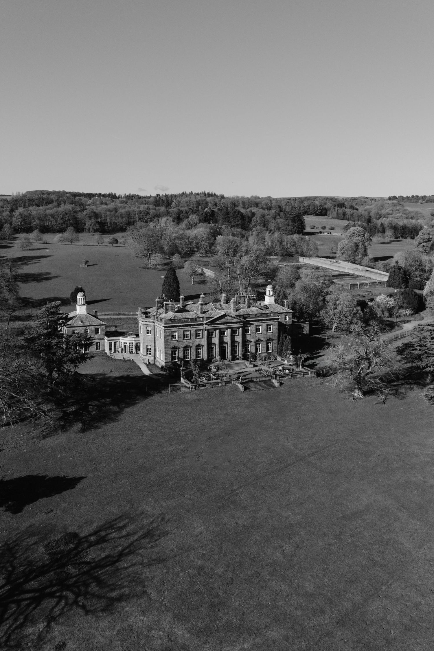 Aerial black and white photo of a grand historic country estate surrounded by lush trees and expansive green lawns.