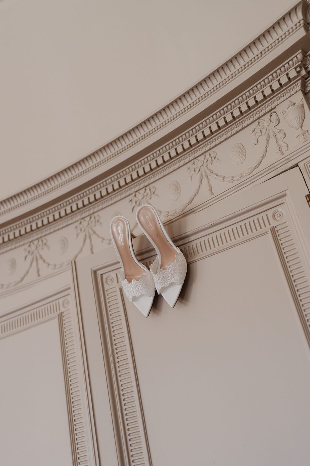 Elegant white bridal heels with lace bow detail displayed on ornate cream molding wall decor.