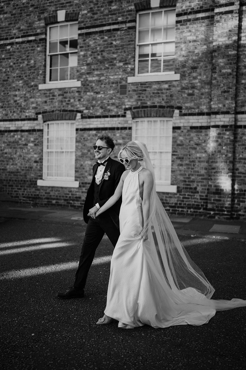 A black and white wedding photo of a couple walking together against a brick building, with the bride in a flowing gown and veil.
