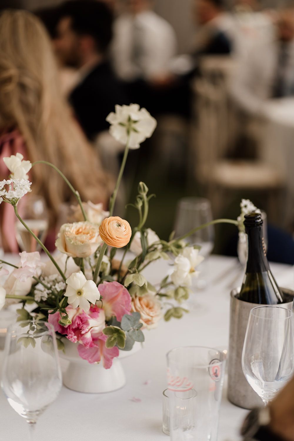 Minimal bright wedding flowers with crockery and wine glasses placed next to them on a white table cloth.