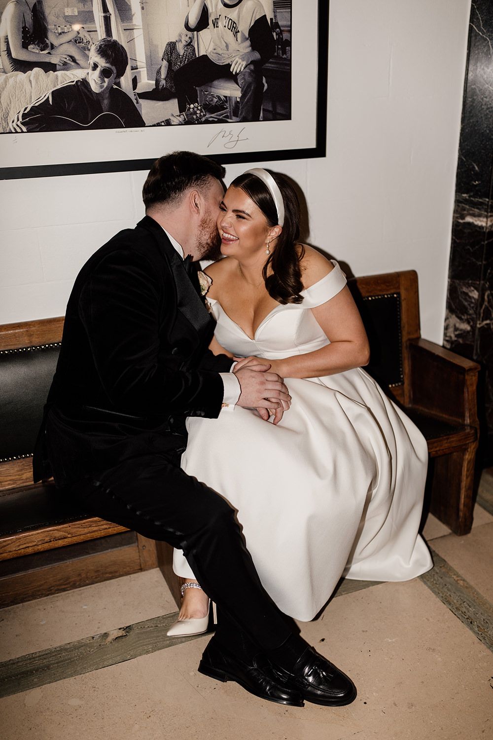 Romantic wedding moment of couple sitting together on wooden bench beneath framed artwork.