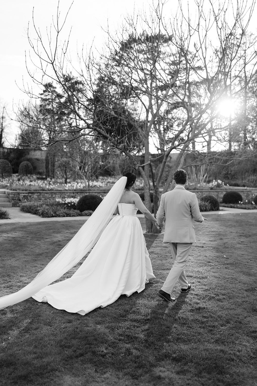 Black and white photo of a couple in wedding attire running playfully through a garden with sunlight peeking through trees.