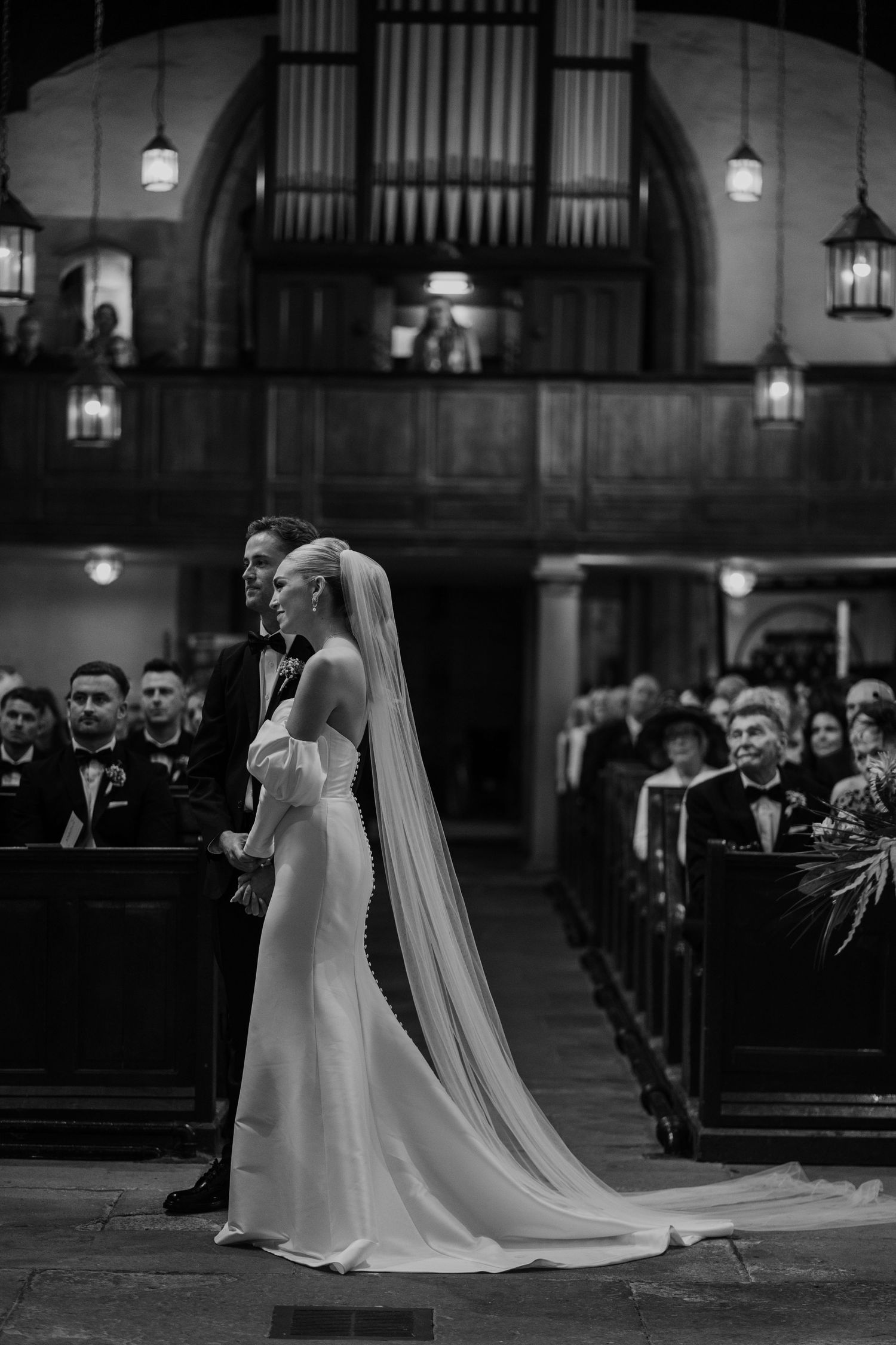 Black and white photograph of a wedding ceremony inside a grand church with organ pipes and hanging lanterns.