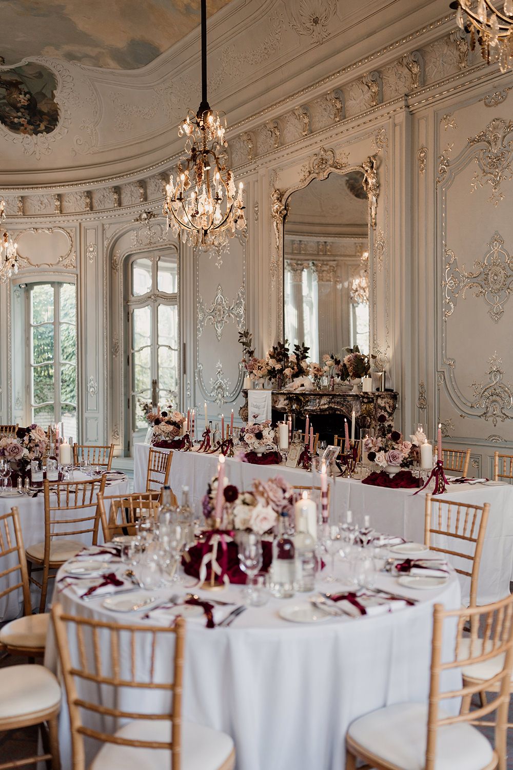 Elegant wedding reception setup in ornate French ballroom with crystal chandeliers and white tables with burgundy accents.