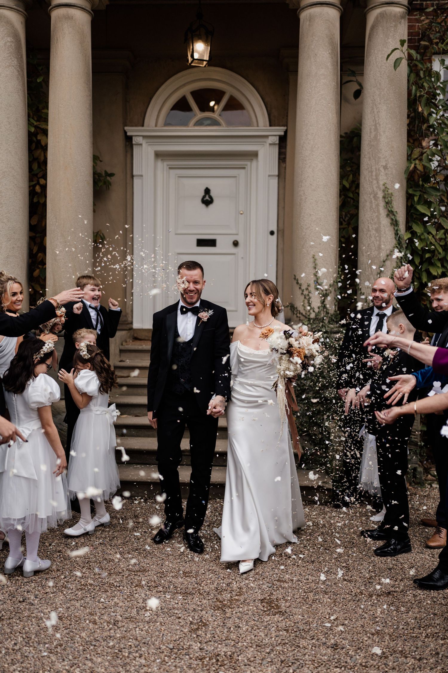 A joyous wedding celebration with guests throwing confetti outside an elegant white-columned entrance with arched doorway.