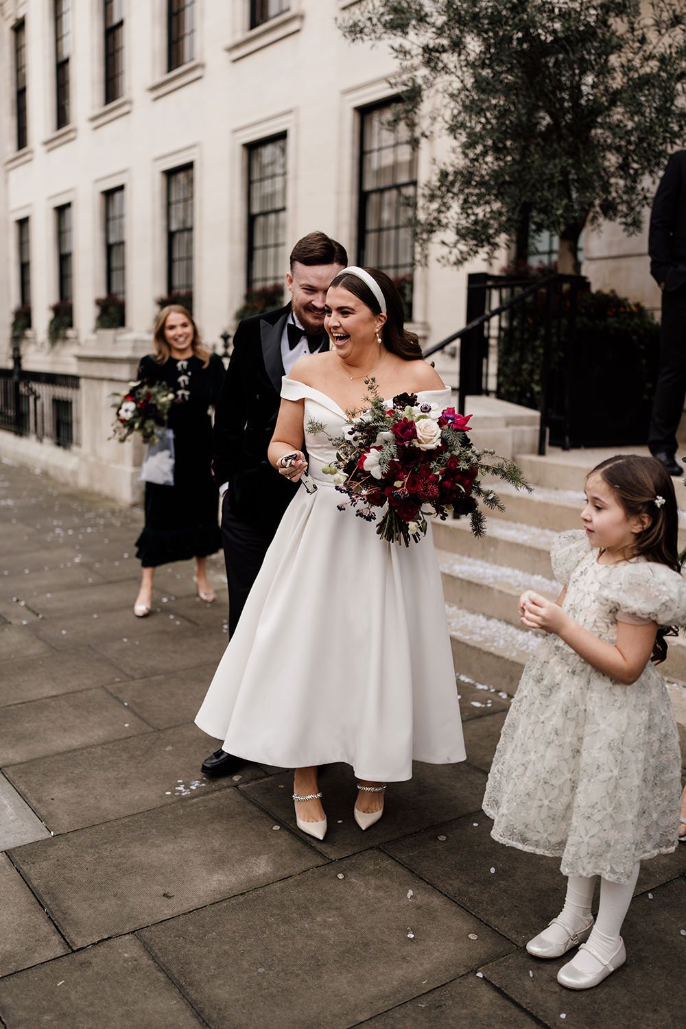 A wedding party with a bride in a tea-length dress and white headband holding a burgundy bouquet on stone church steps.