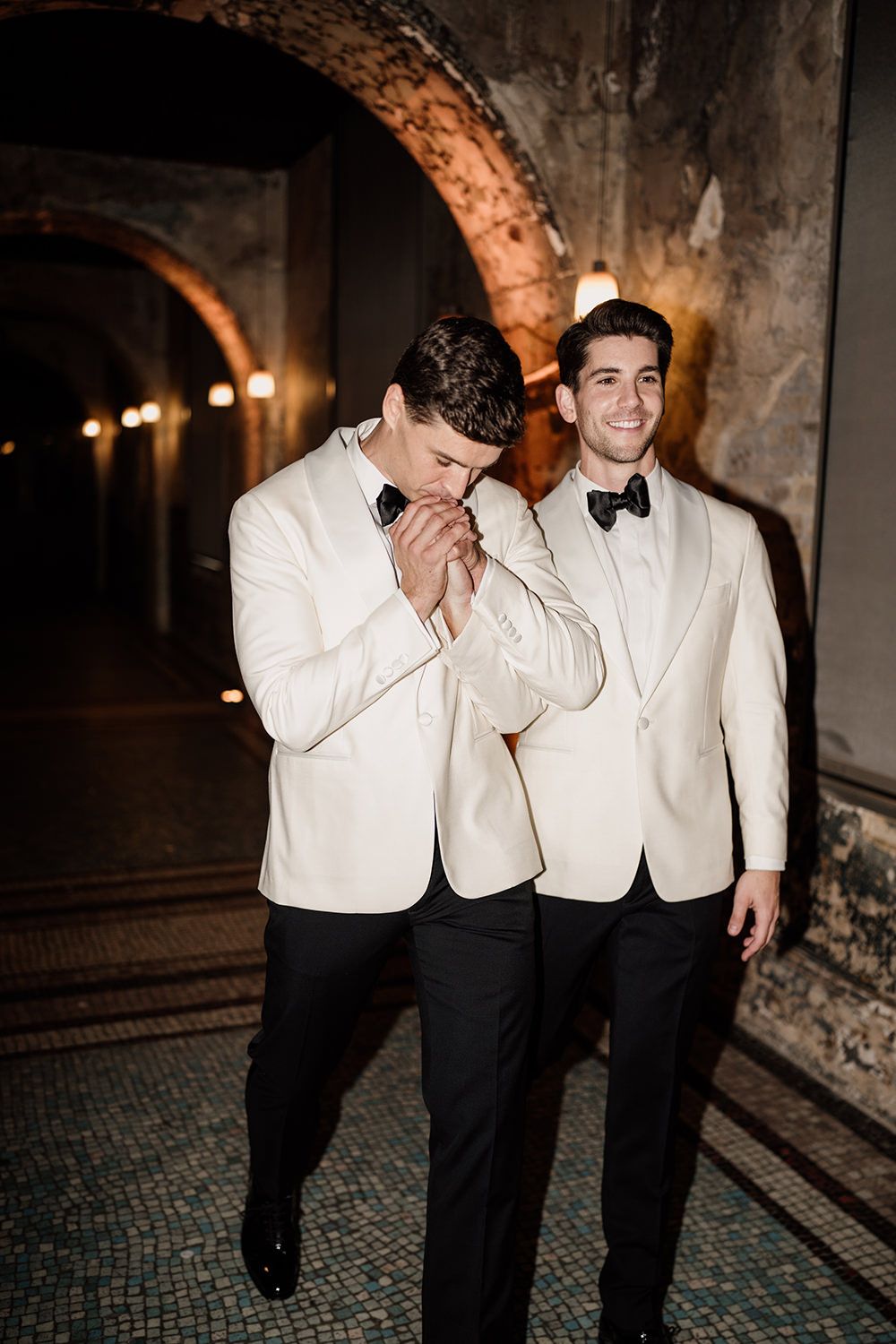 Grooms walk laughing in white and black suits having a photo taken with a harsh flash.