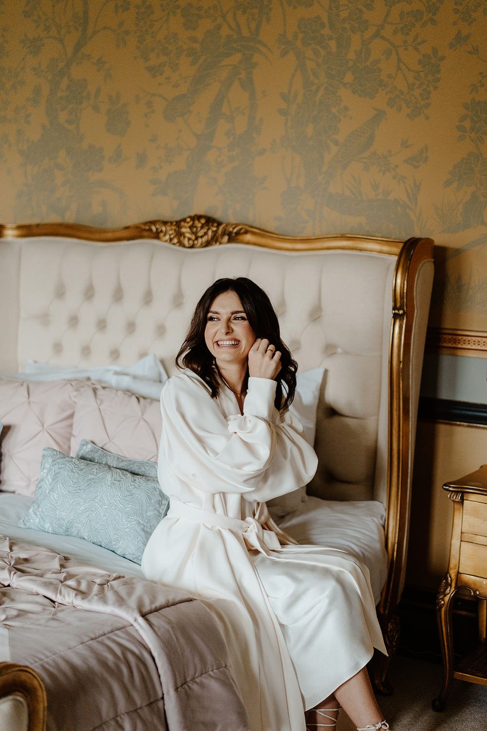 Bride sat on a grand bed while she puts in her wedding earrings.