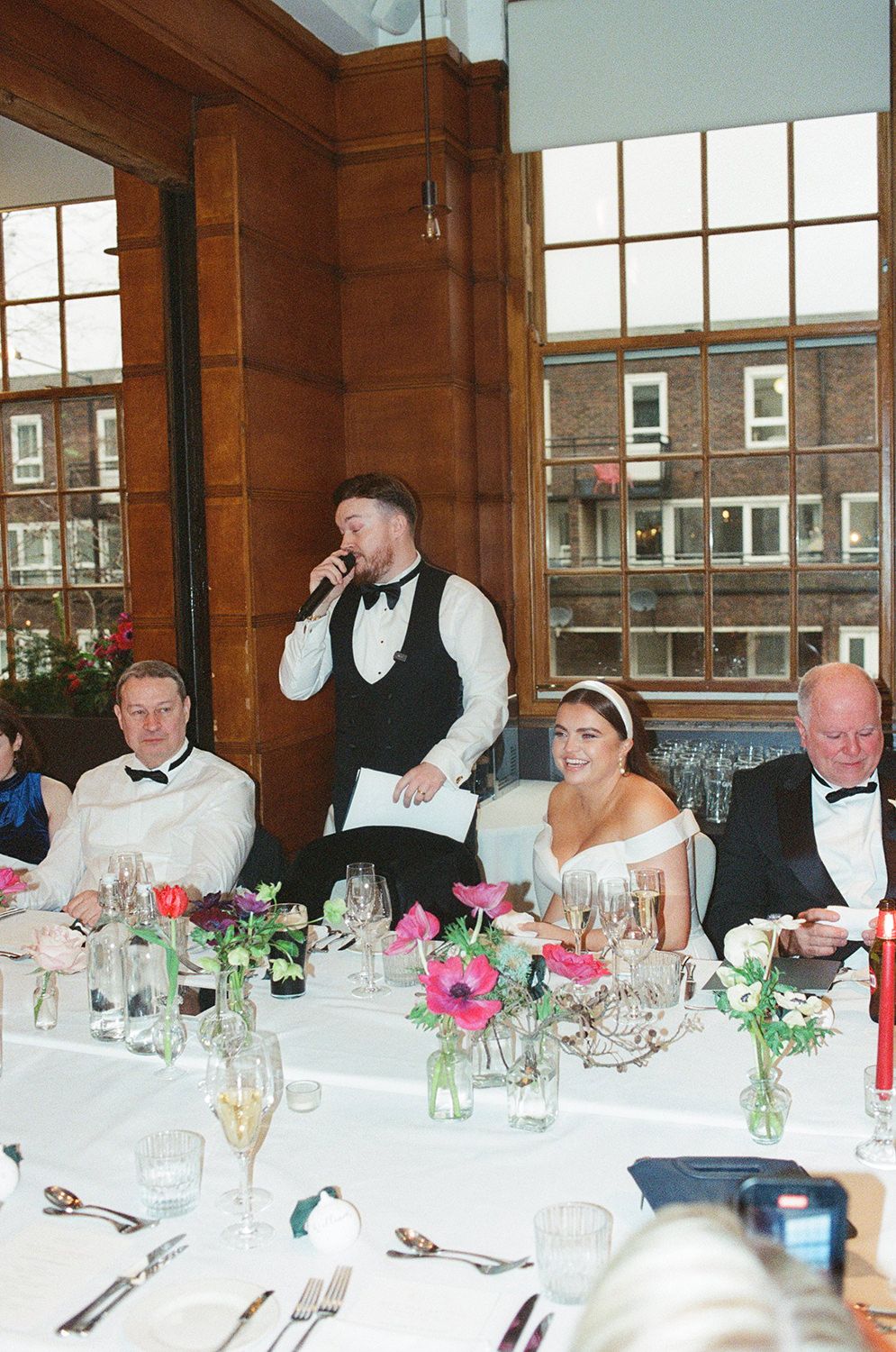 Wedding reception scene with table settings decorated with pink flowers and guests seated for dinner.