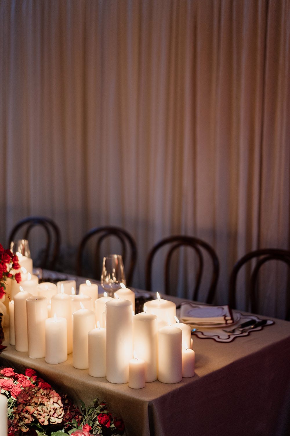 White pillar candles line a dark wooden table, creating a romantic glow beside delicate red floral arrangements.
