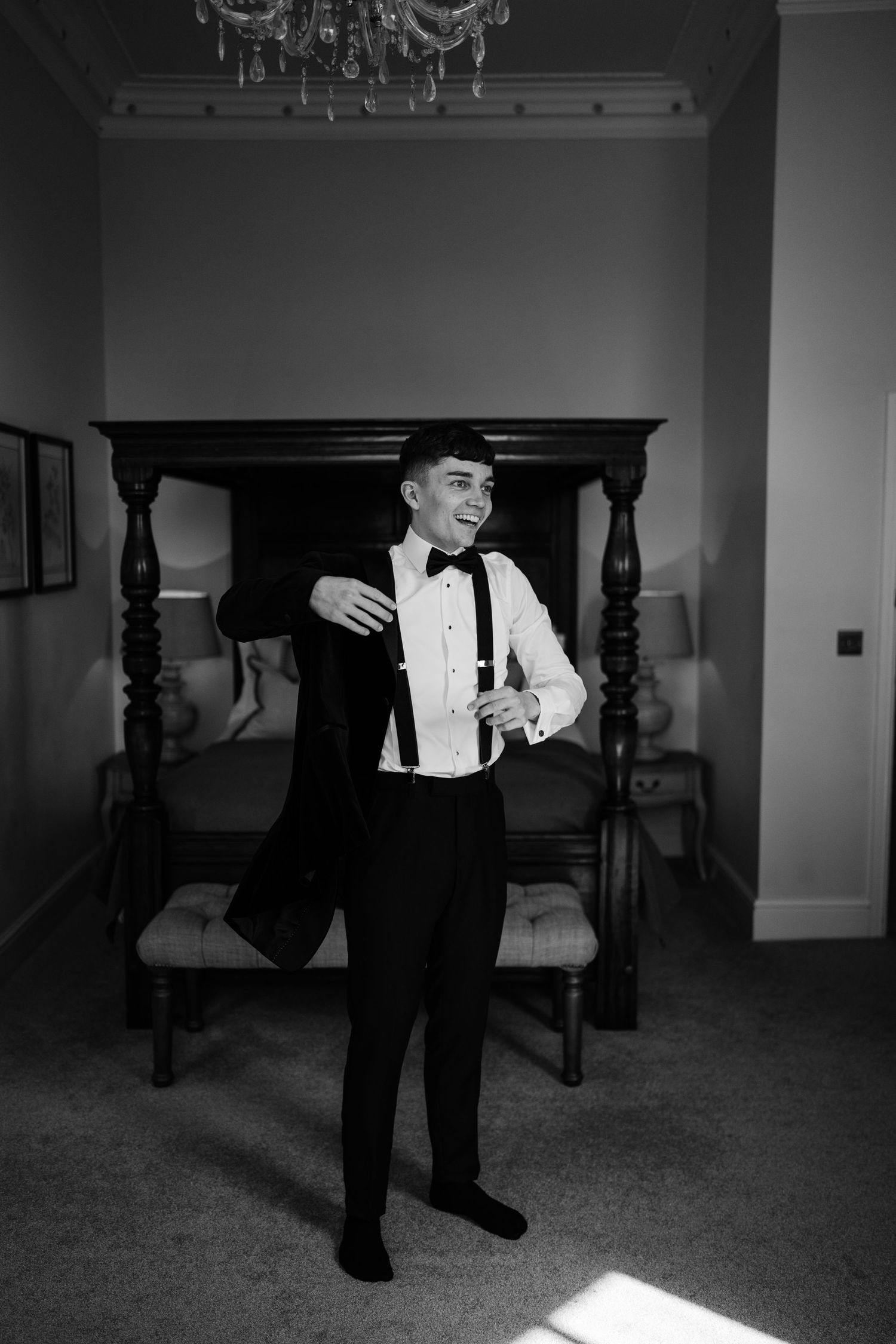 Groom putting on his blazer with a wooden bed in the background while smiling.