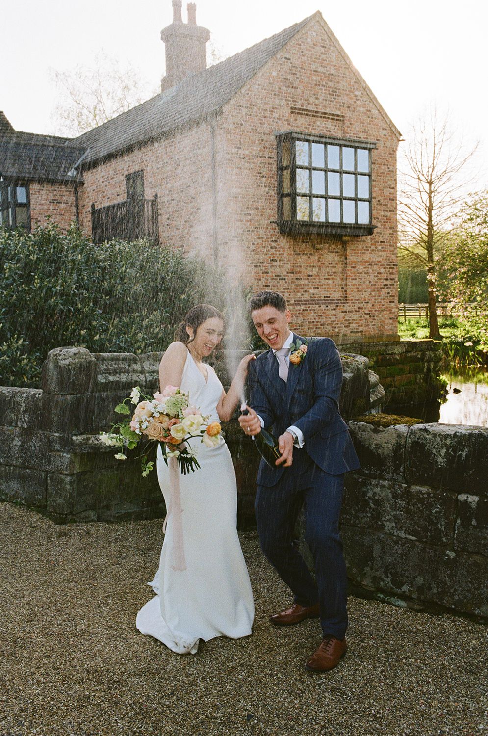 Bride and groom smiling and cheering while spraying champagne with Holford Hall behind them.
