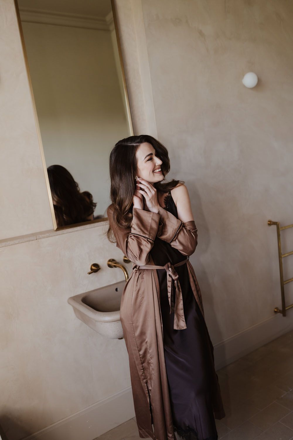 A smiling woman in a satin bronze robe stands in an elegant bathroom near a white sink and mirror.