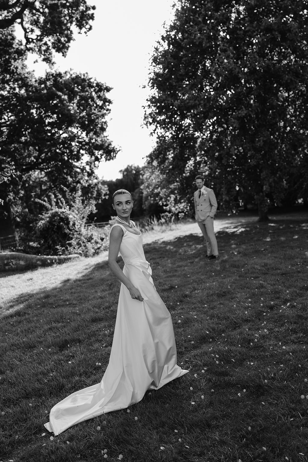 Black and white wedding photo of bride in flowing gown with groom standing in background among lush garden trees.