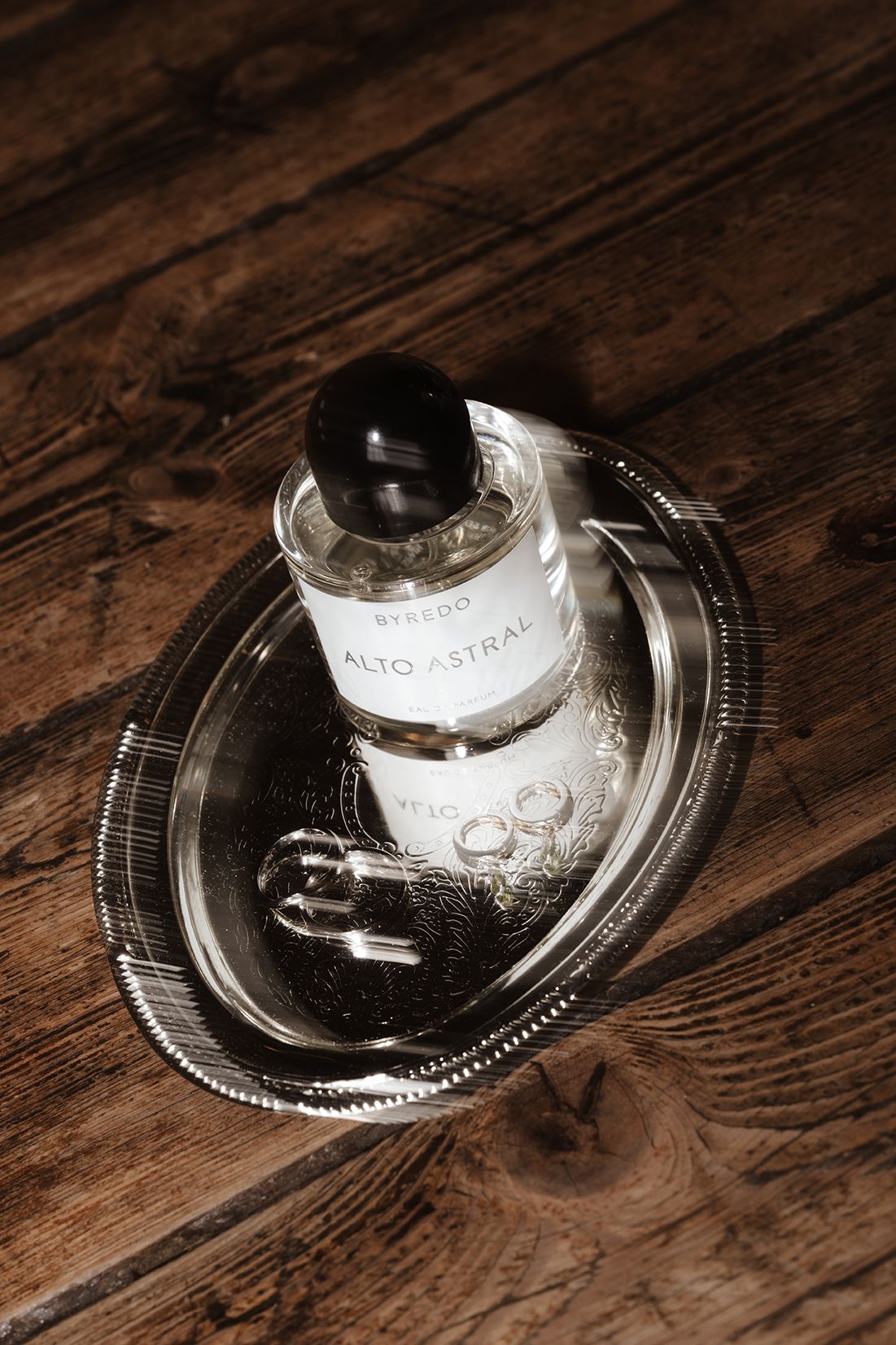 Byredo Alto Astral perfume bottle with wedding rings on a silver tray atop a rustic wood surface.