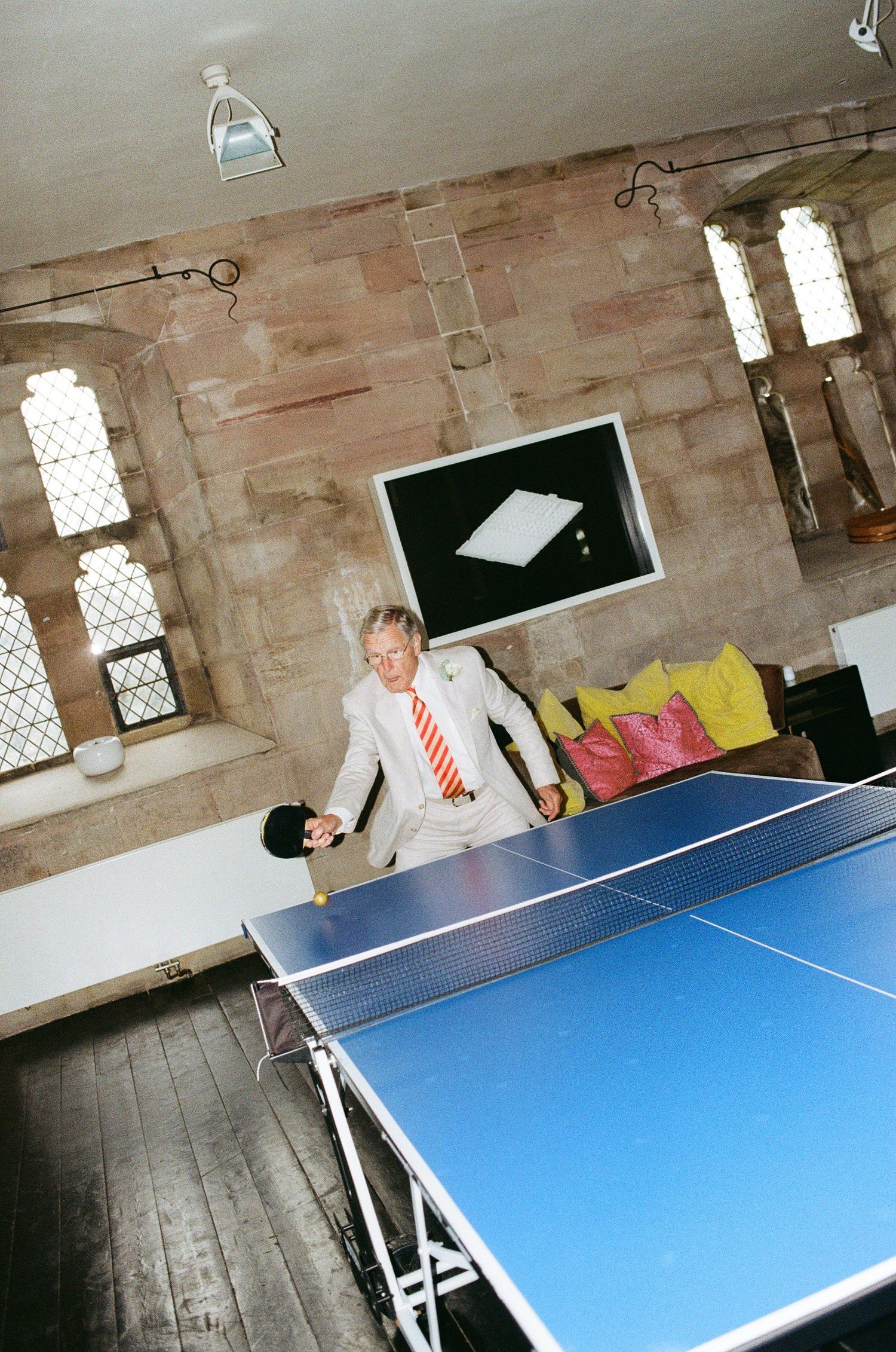 Man in suit playing ping pong on a blue table tennis table inside a rustic stone-walled room.