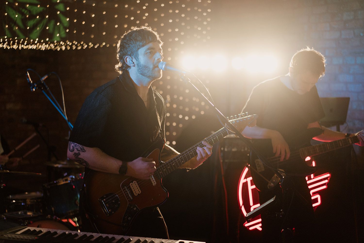 Guitarist stood playing his guitar while singing into a microphone with stage lights bright behind him.