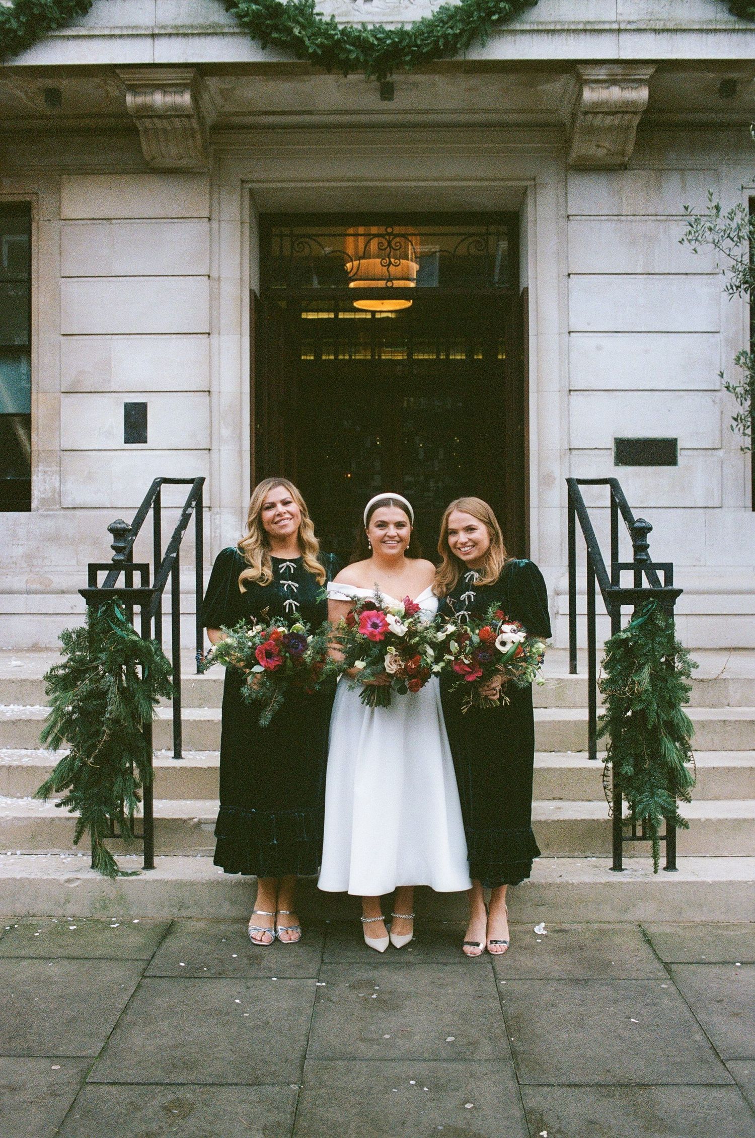 Three figures in black and white attire stand on stone steps of an elegant building decorated with holiday greenery.