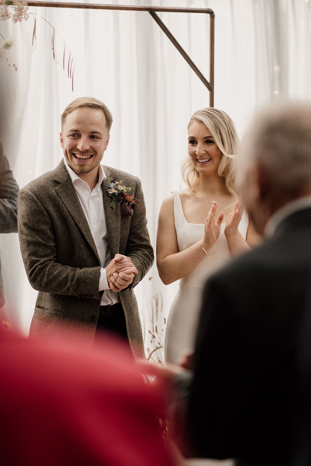 Bride and groom stood smiling and clapping in-front of wedding guests.
