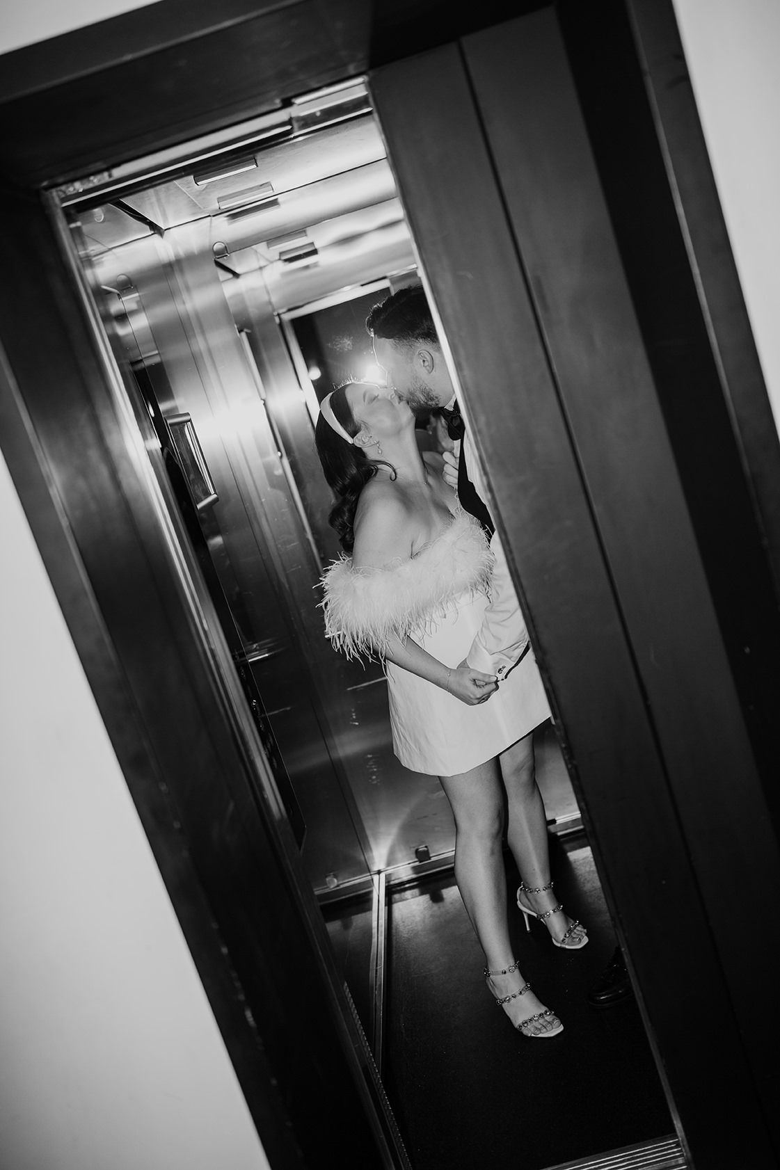 A black and white photograph shows a figure in a feathered dress standing in an elevator.