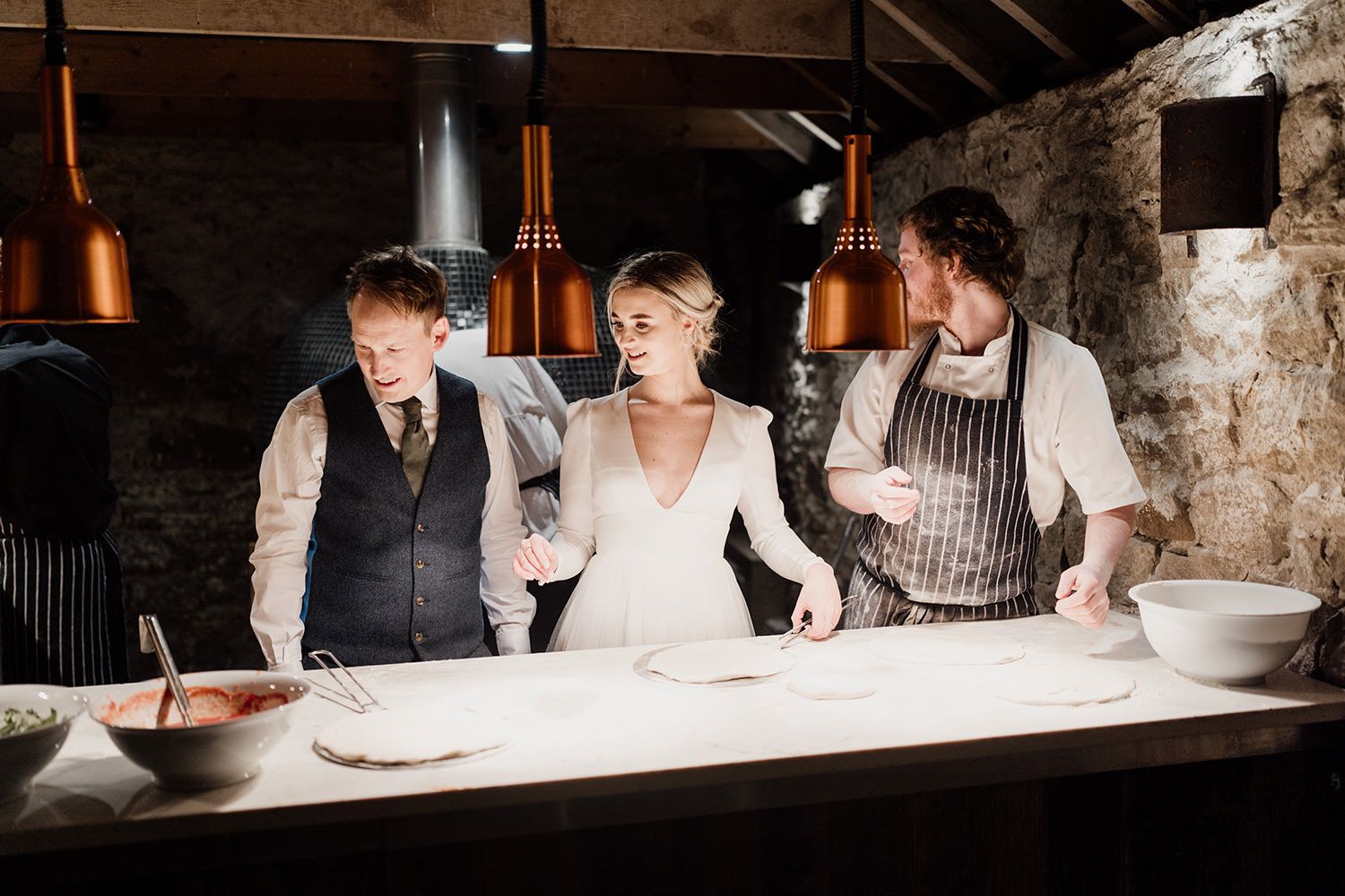 Bride and groom stood being shown how to make pizza on a white table.