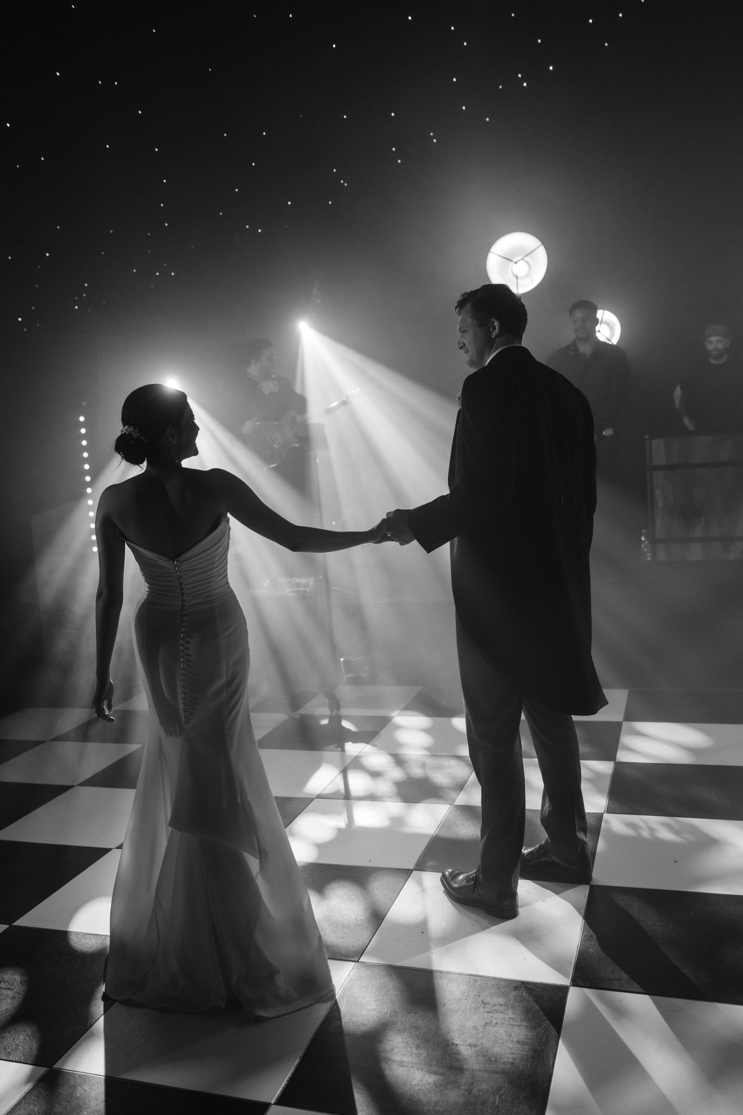 Bride and groom share first dance on checkered floor under dramatic spotlight in black and white wedding photo.