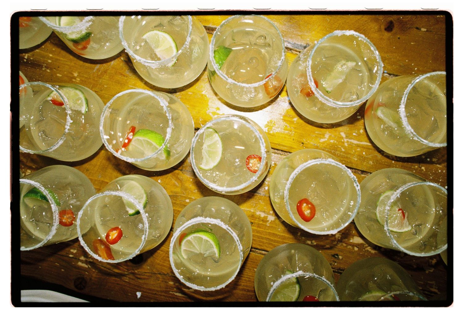 Multiple margarita cocktails with lime slices and chili peppers on salted rims arranged on a wooden table.
