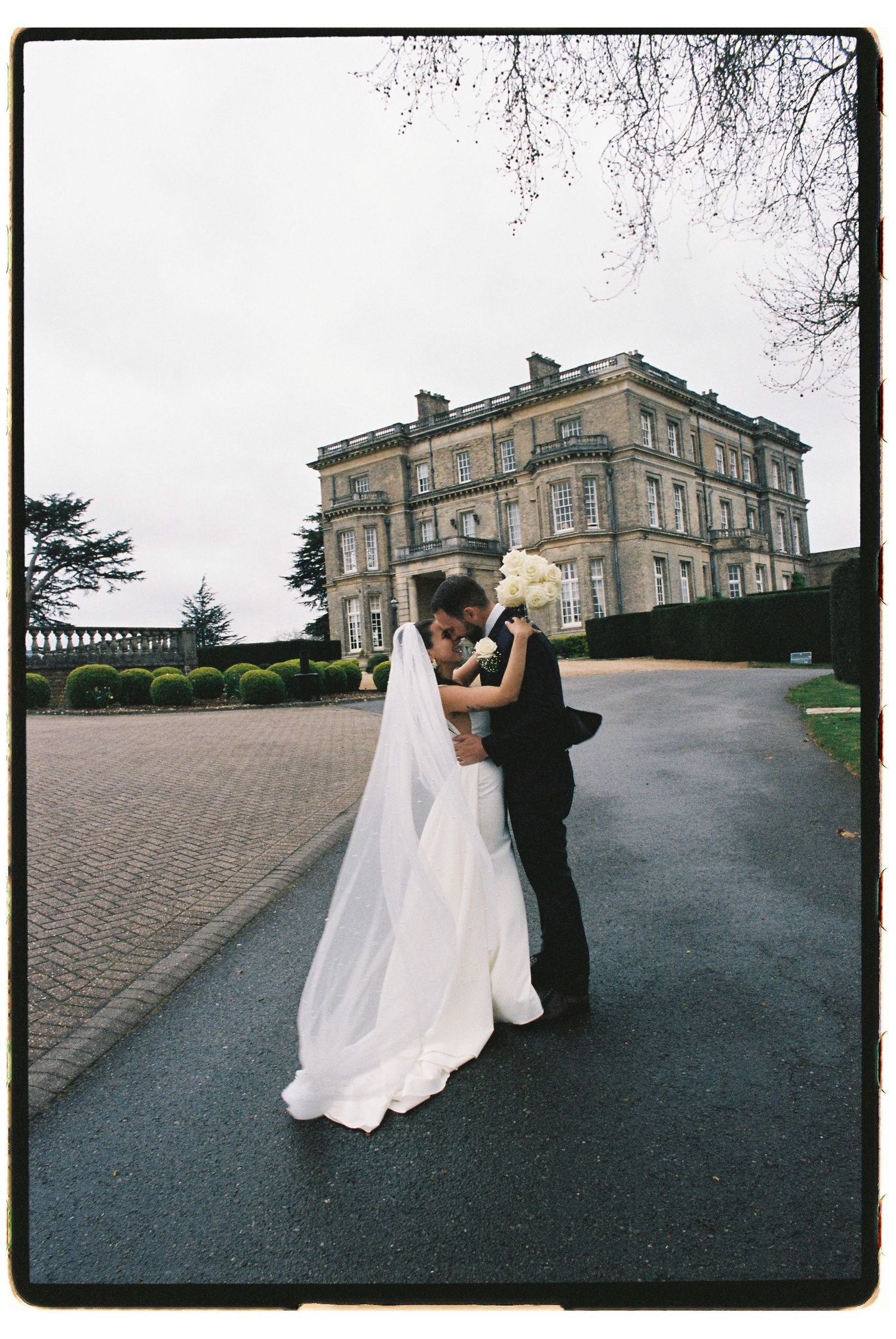 Bride and groom embracing in front of a grand historic mansion on their wedding day, bride wearing a flowing white gown and veil.
