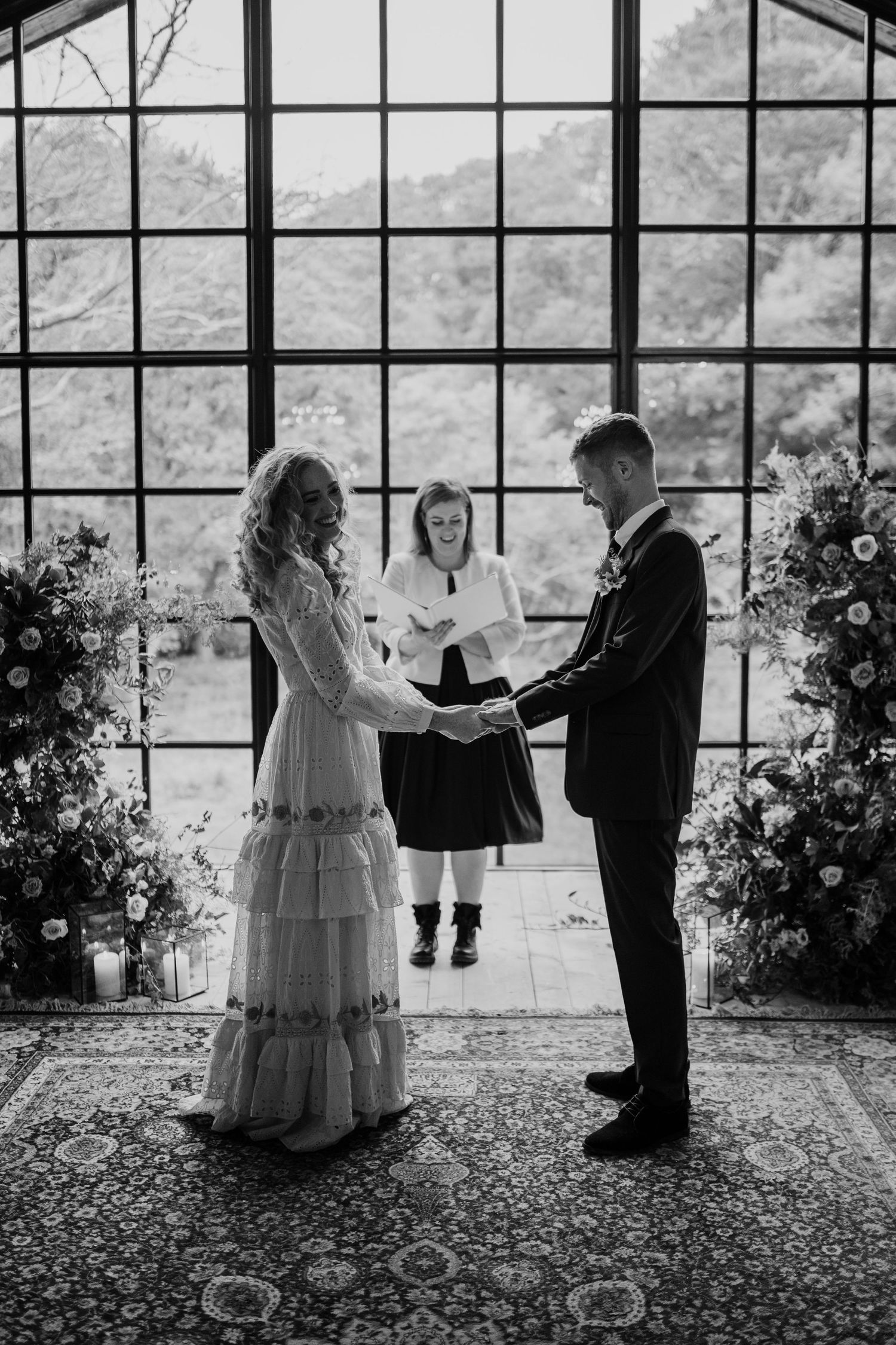 Couple holding hands during wedding ceremony with barn windows and celebrant behind them.