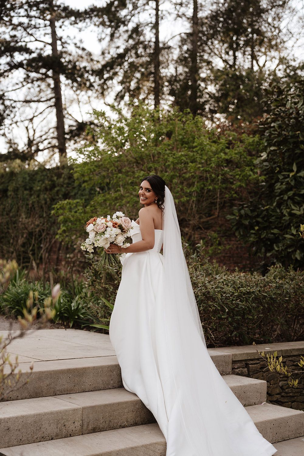 A person in an elegant white wedding gown stands on concrete steps holding a white floral bouquet in a garden setting.