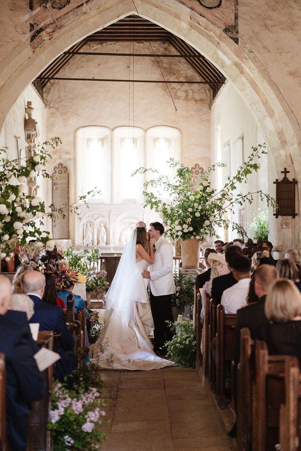A romantic church wedding ceremony with beautiful floral decorations and guests seated in wooden pews under an arched ceiling.