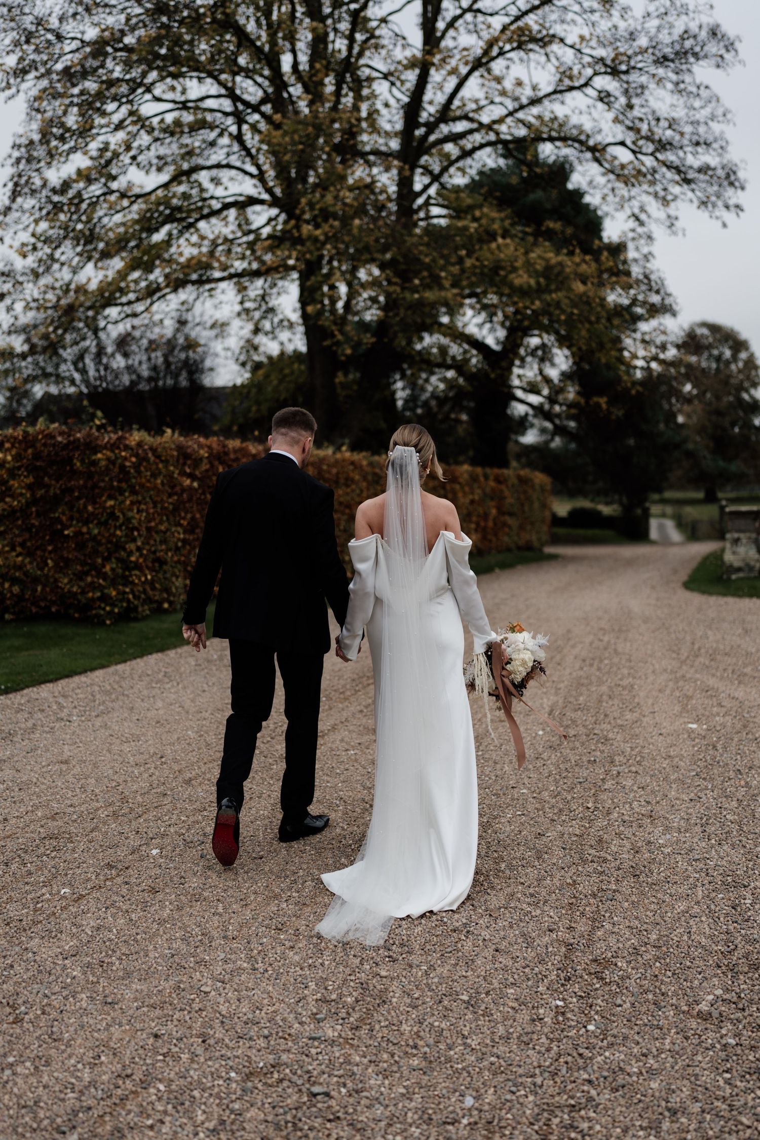 A couple walks down a gravel path in a garden setting, with the bride wearing a flowing white dress and holding a bouquet.
