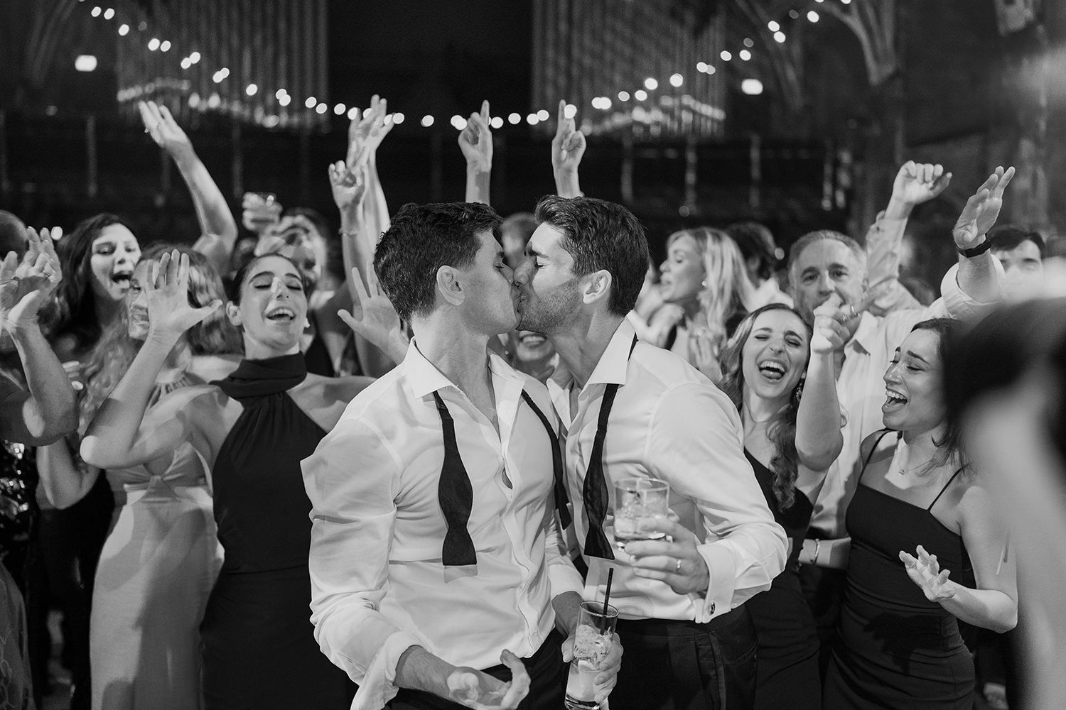 Two grooms kissing at the end of their wedding night with guests around them celebrating.