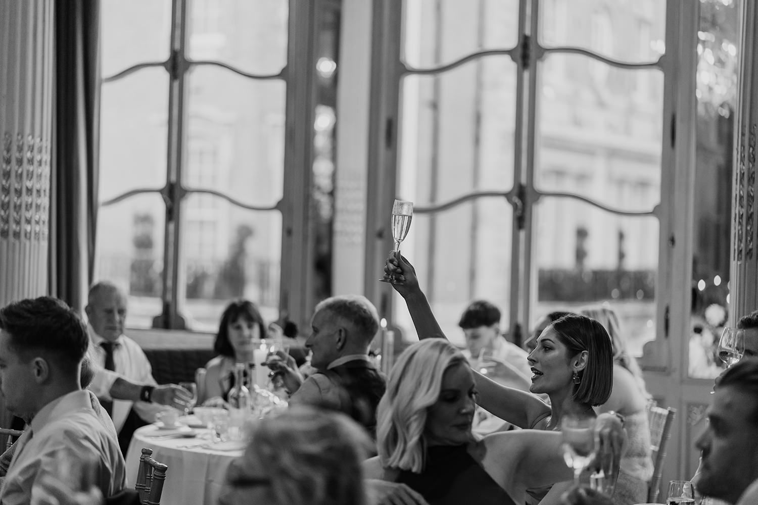 Wedding reception guests seated at long tables in bright sunlit venue with large windows.