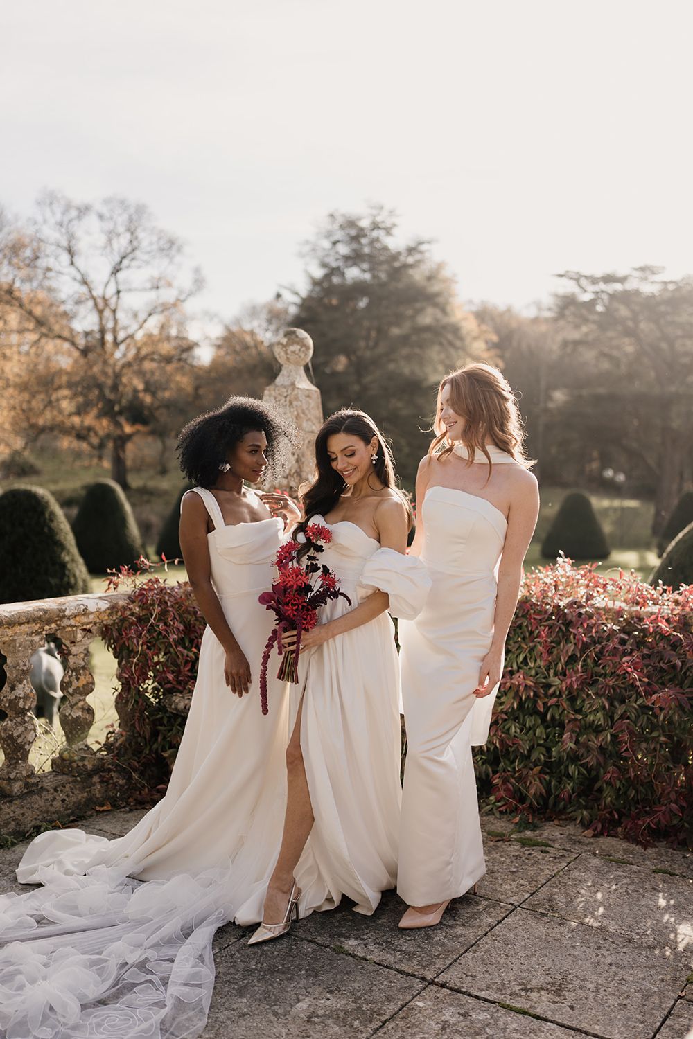 A group wearing white dresses gathered on a garden terrace at sunset with floral bouquets and soft sunlight filtering through trees.