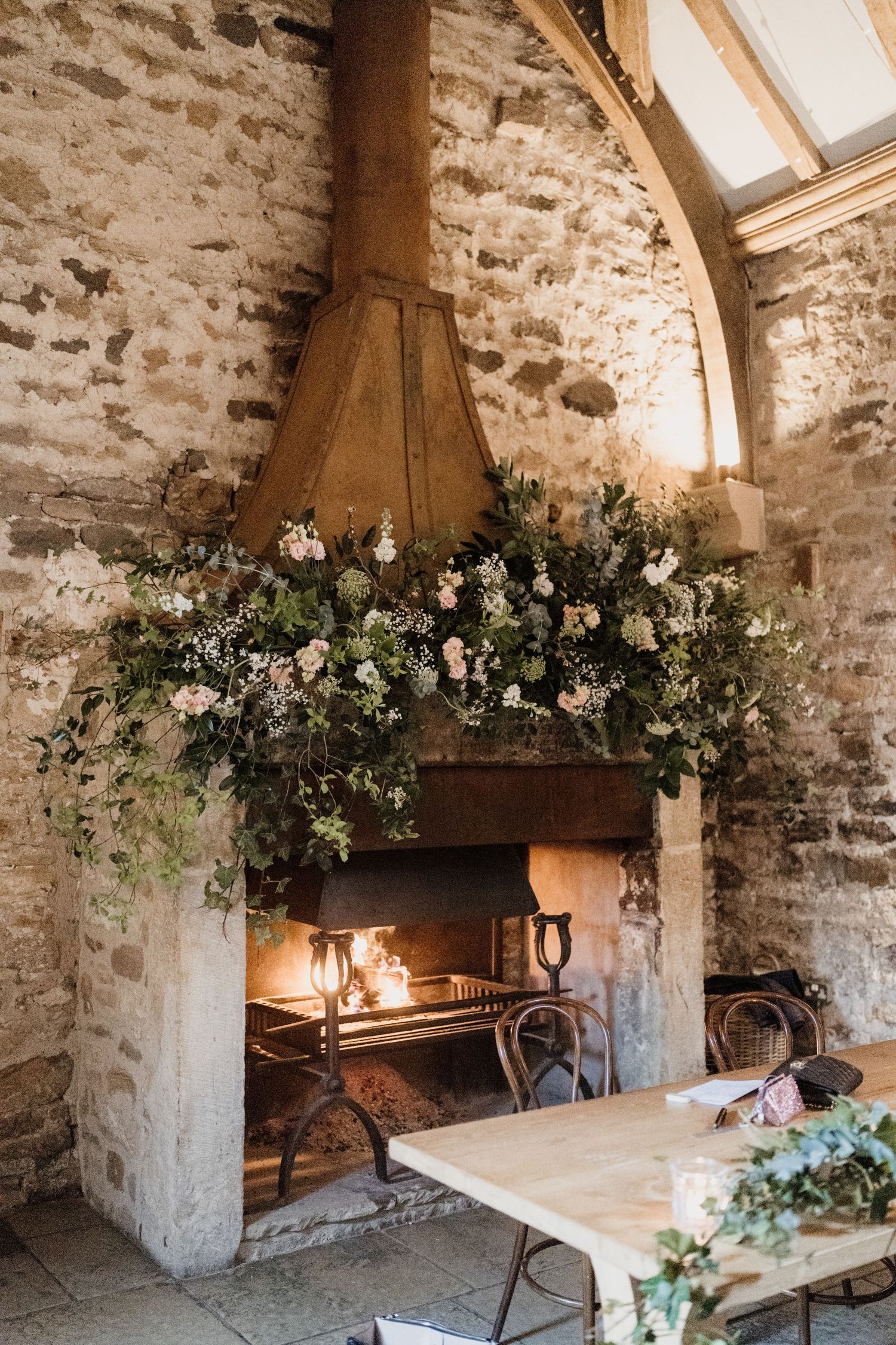 A rustic stone wall interior decorated with lush floral arrangements and greenery garlands around wooden beams and fixtures.