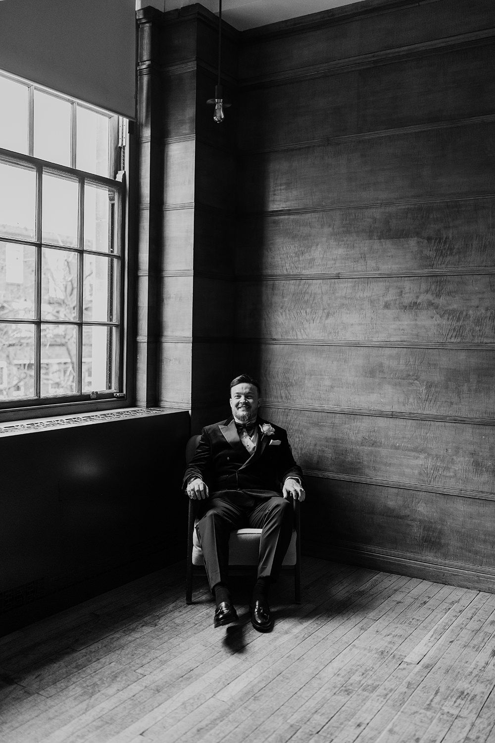 Black and white photograph of someone seated in a wooden chair against a wall near a large window with natural light.