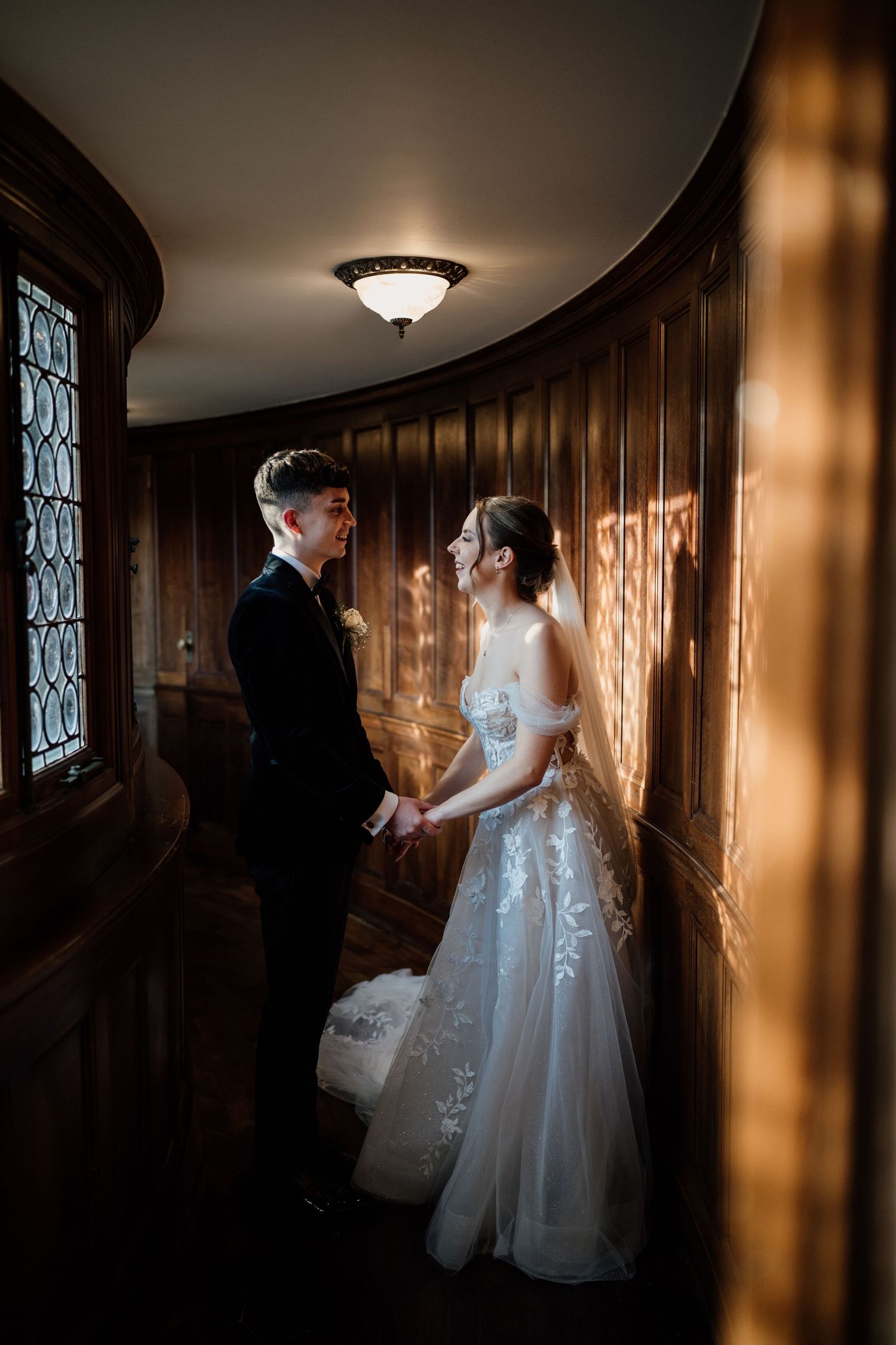 Bride and groom smiling facing towards eachother as light streams through a window in a wooden corridor at Bourton Hall.