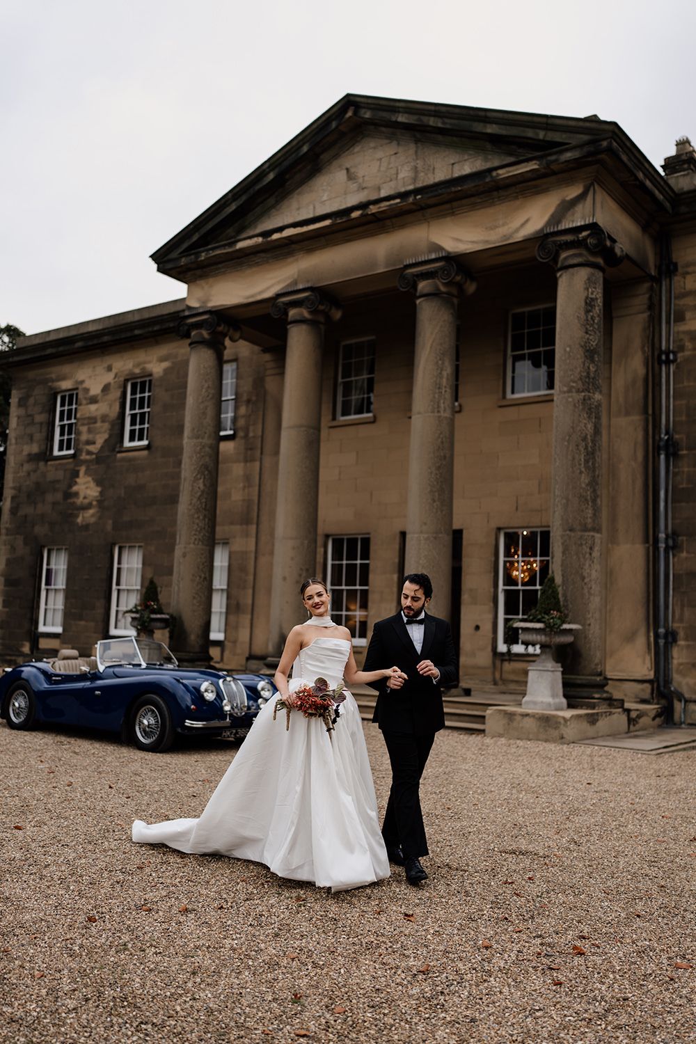 Bride and groom laughing and running through the grounds at Rise Hall with a vintage car in the background.