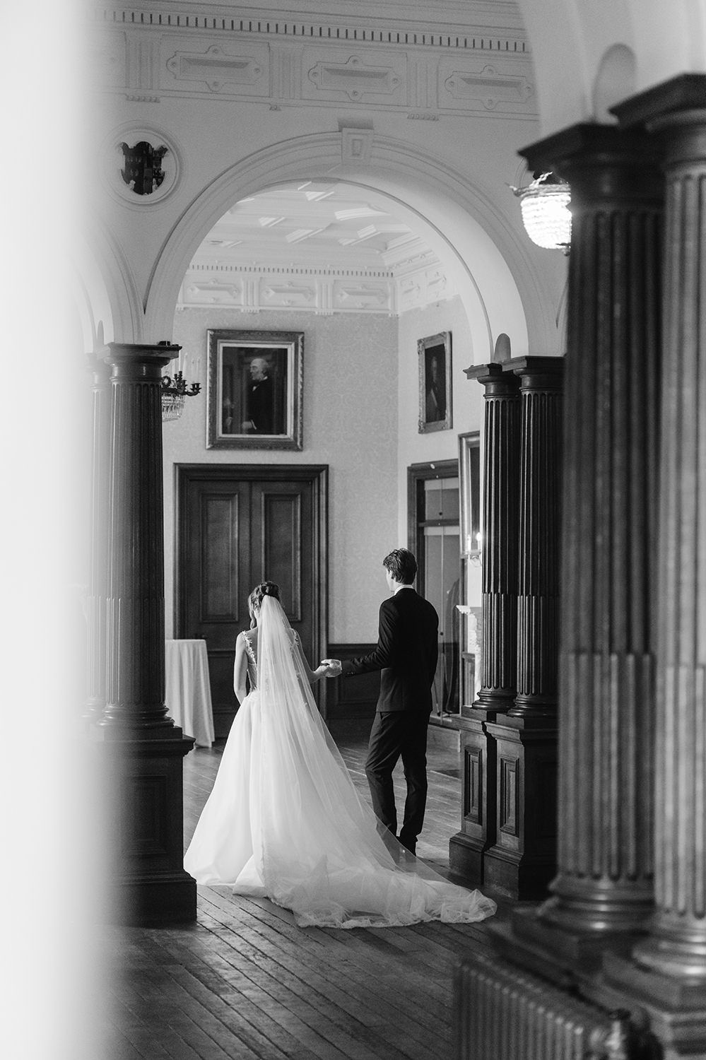 A bride and groom walk through an elegant hallway with classical columns and white walls in black and white.