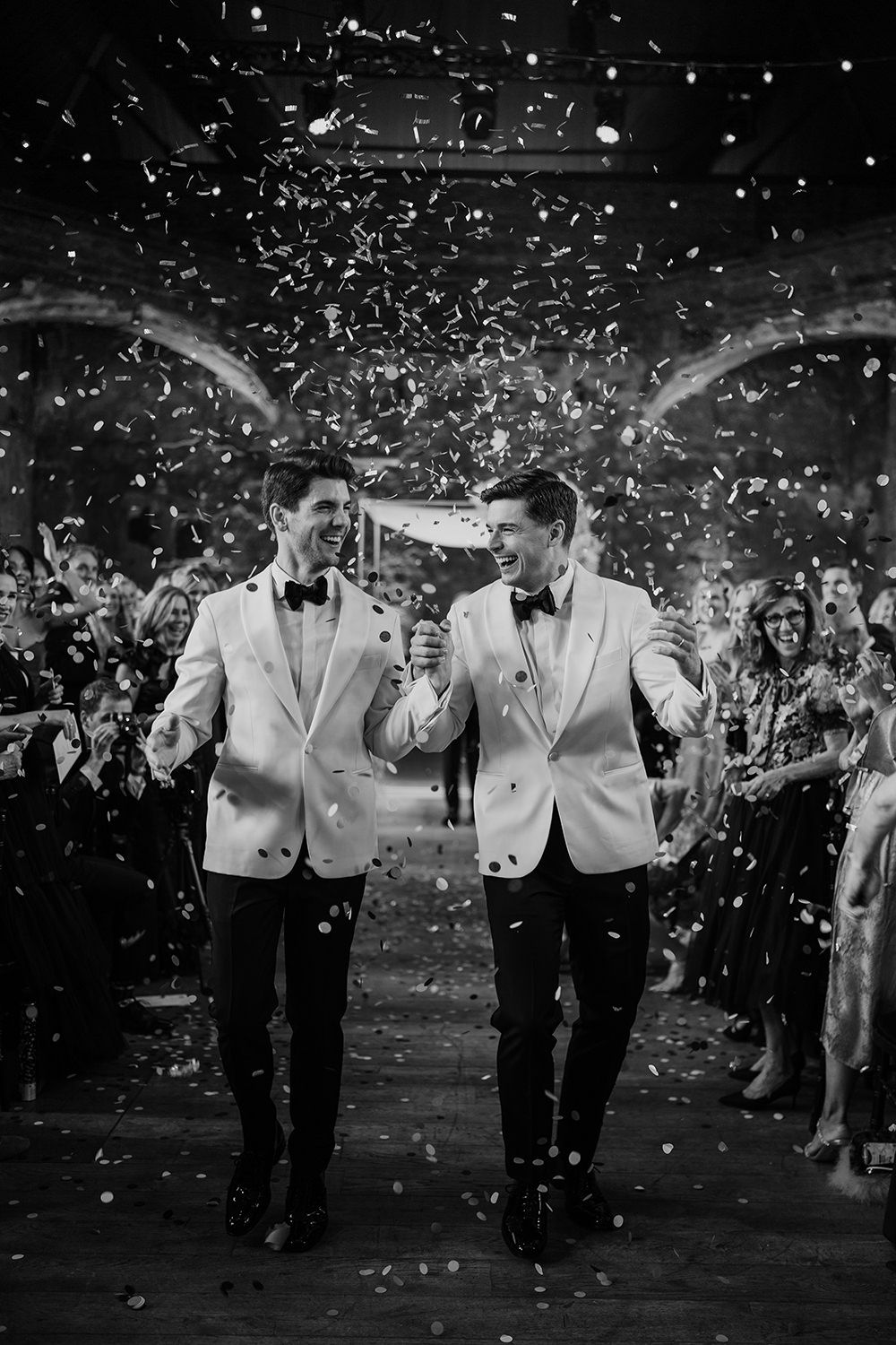 Two grooms walking through silver confetti laughing and celebrating with guests.